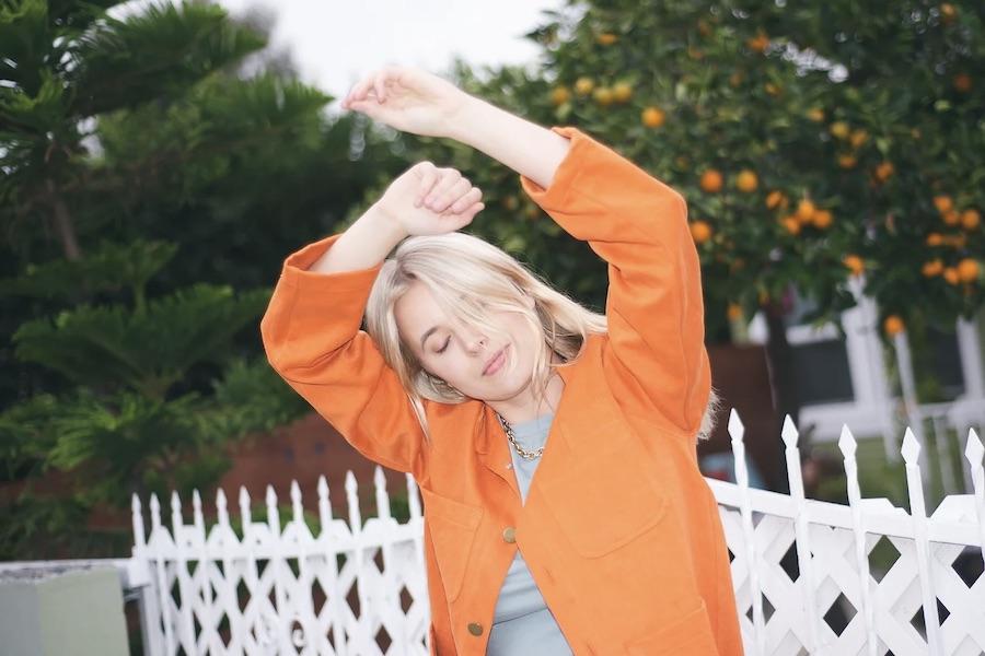 A young woman in a bright orange jacket closes her eyes and waves her arms over her head. Behind her is a white picket fence and a tree with oranges.