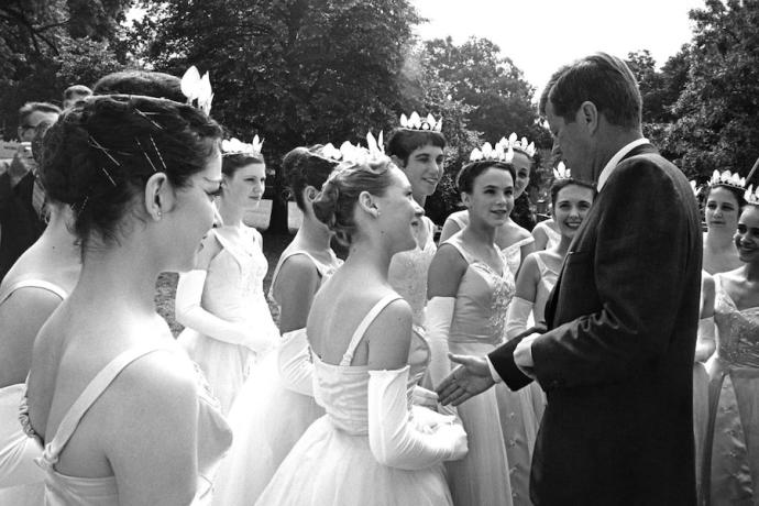 A black-and-white photo of a group of young dancers shaking hands with President John F. Kennedy.